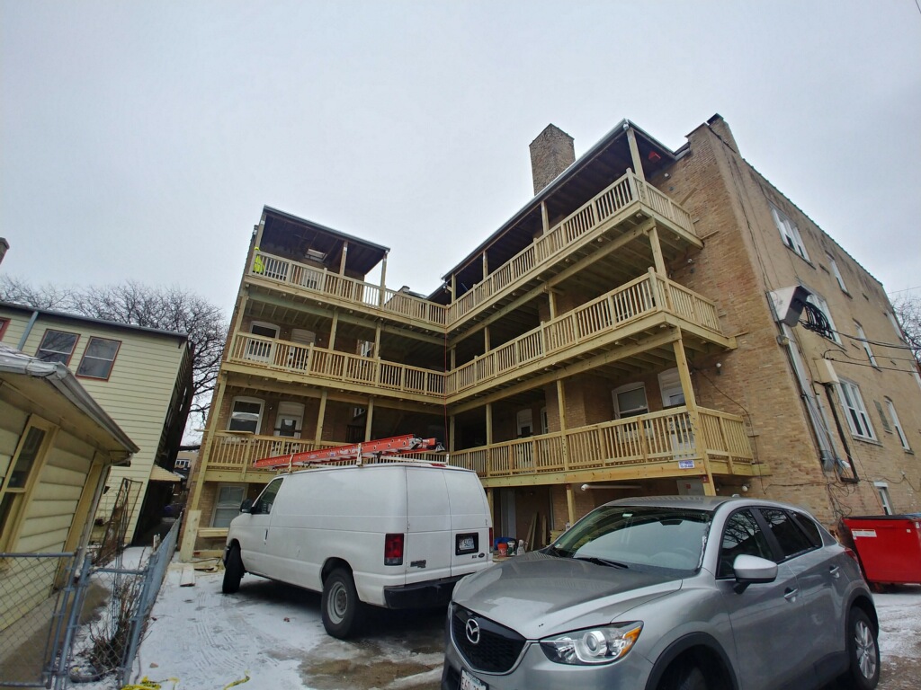 a multi-level building with wooden balconies and staircases, vehicles parked in front.