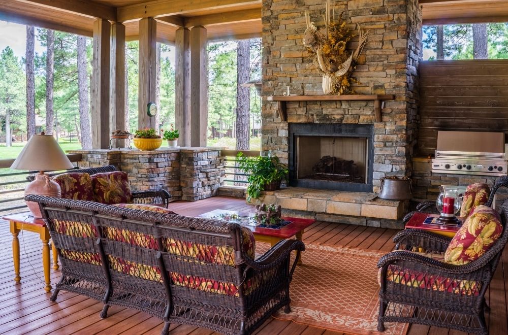 A rustic Porch area with a stone fireplace, wicker furniture with patterned cushions, and a view of pine trees.