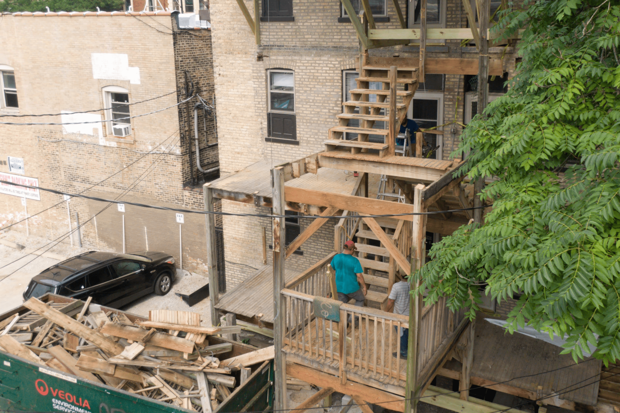 A multi-level wooden fire escape on a brick building with people walking on the stairs, adjacent to a leafy tree- chicgao porch repair with city porches