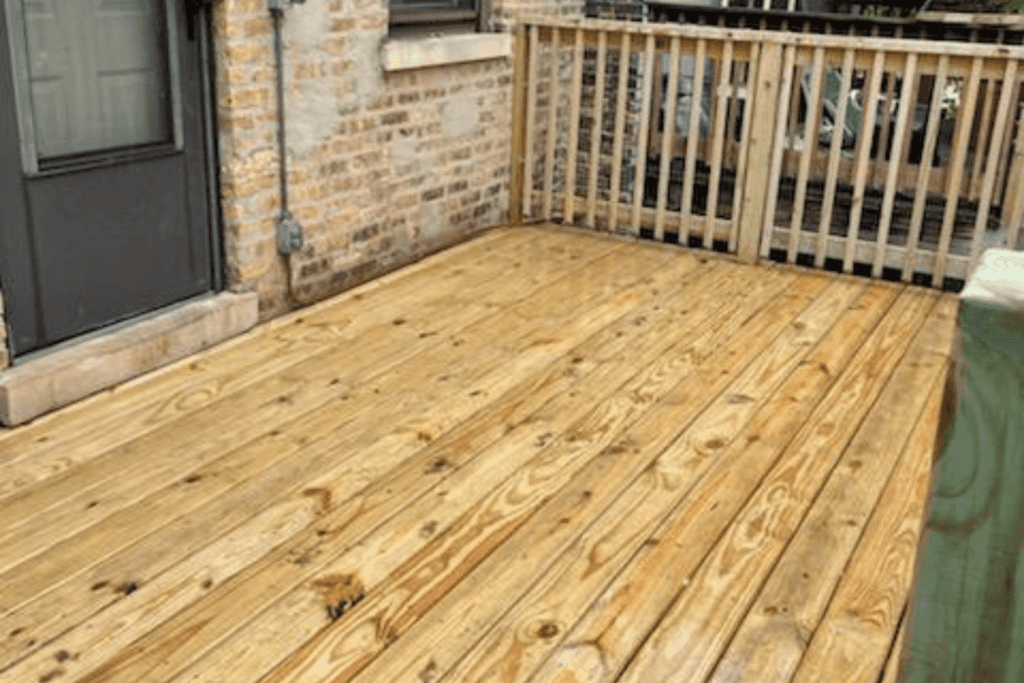 Wooden deck flooring with a detailed grain pattern near a building's brick wall and a railing.- Our services