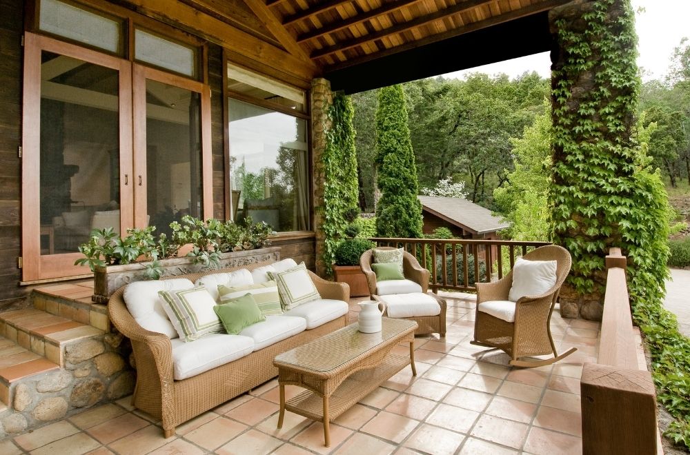 An inviting covered patio with wicker furniture, white cushions, terracotta tiles, and lush greenery climbing the pillars.