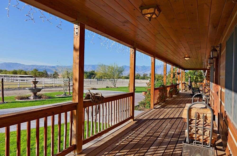 A long wooden porch with rustic railings and furniture, overlooking a fenced yard and mountains in the distance.