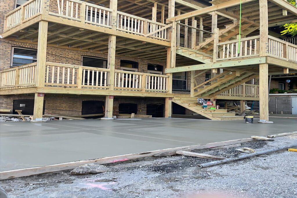 Freshly poured concrete foundation in front of a multi-story building with new wooden porches and staircases under construction.-City Porches Roscoe Village | Porch and deck repair in Chicago - Cityporches.com