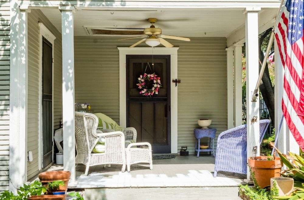 A welcoming front entry with wicker furniture, a ceiling fan, floral wreath on the door, and an American flag.