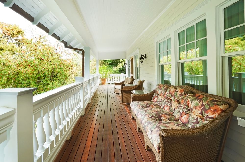 A spacious porch with wicker furniture, floral cushions, white railing, greenery outside, and a classic white exterior.