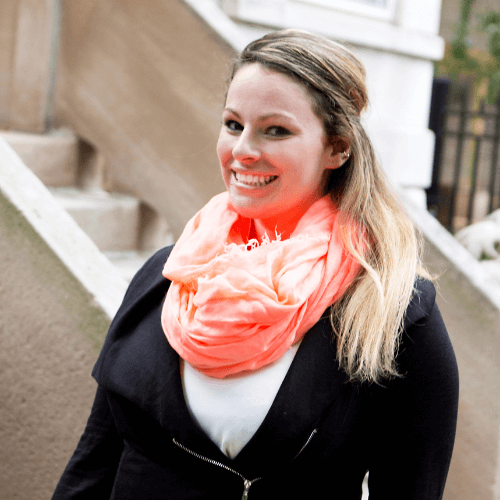 A smiling woman with blonde hair tied back, wearing a black coat and a bright coral scarf, standing on outdoor steps.