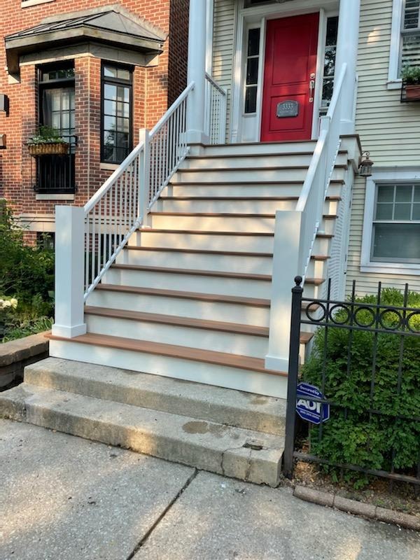 A classic brick house entrance with a striking red door framed by white architectural details. White stairs with handrails lead up to the door, flanked by lush greenery and a small wrought iron fence. A blue sale sign is visible to one side on the ground.