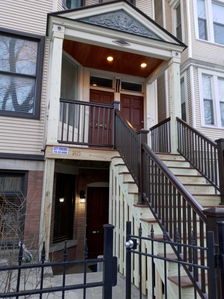 A multi-story residential building entrance with a set of wooden stairs leading to a covered porch with decorative lighting. The porch features a detailed ornamental ceiling design, warm lighting, and a pair of dark wooden doors. A wrought iron fence encloses the front of the property.
