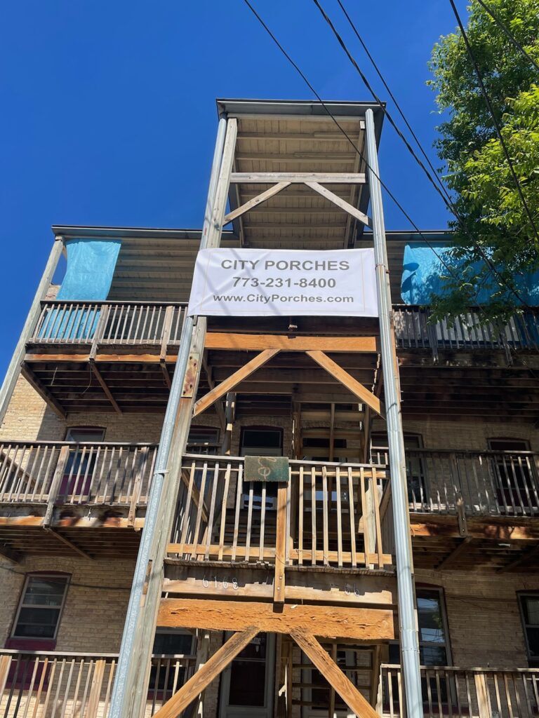 A three-story building with a wooden porch structure under clear blue skies, featuring a banner with contact information.