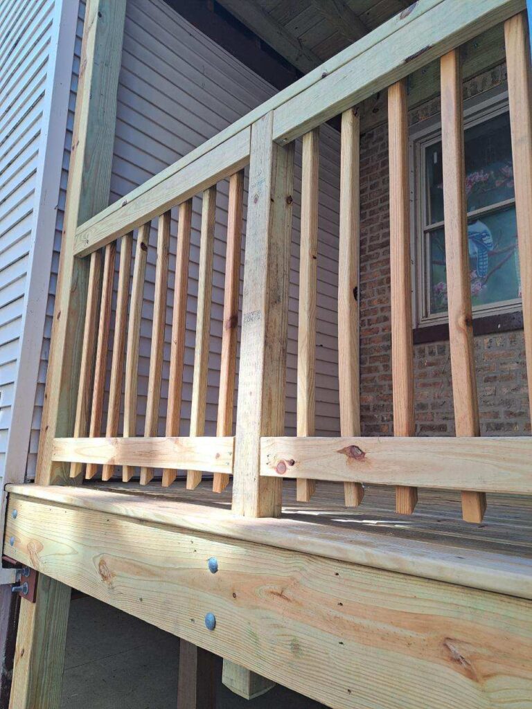 Close-up of a wooden deck railing and posts with a brick building and siding in the background.