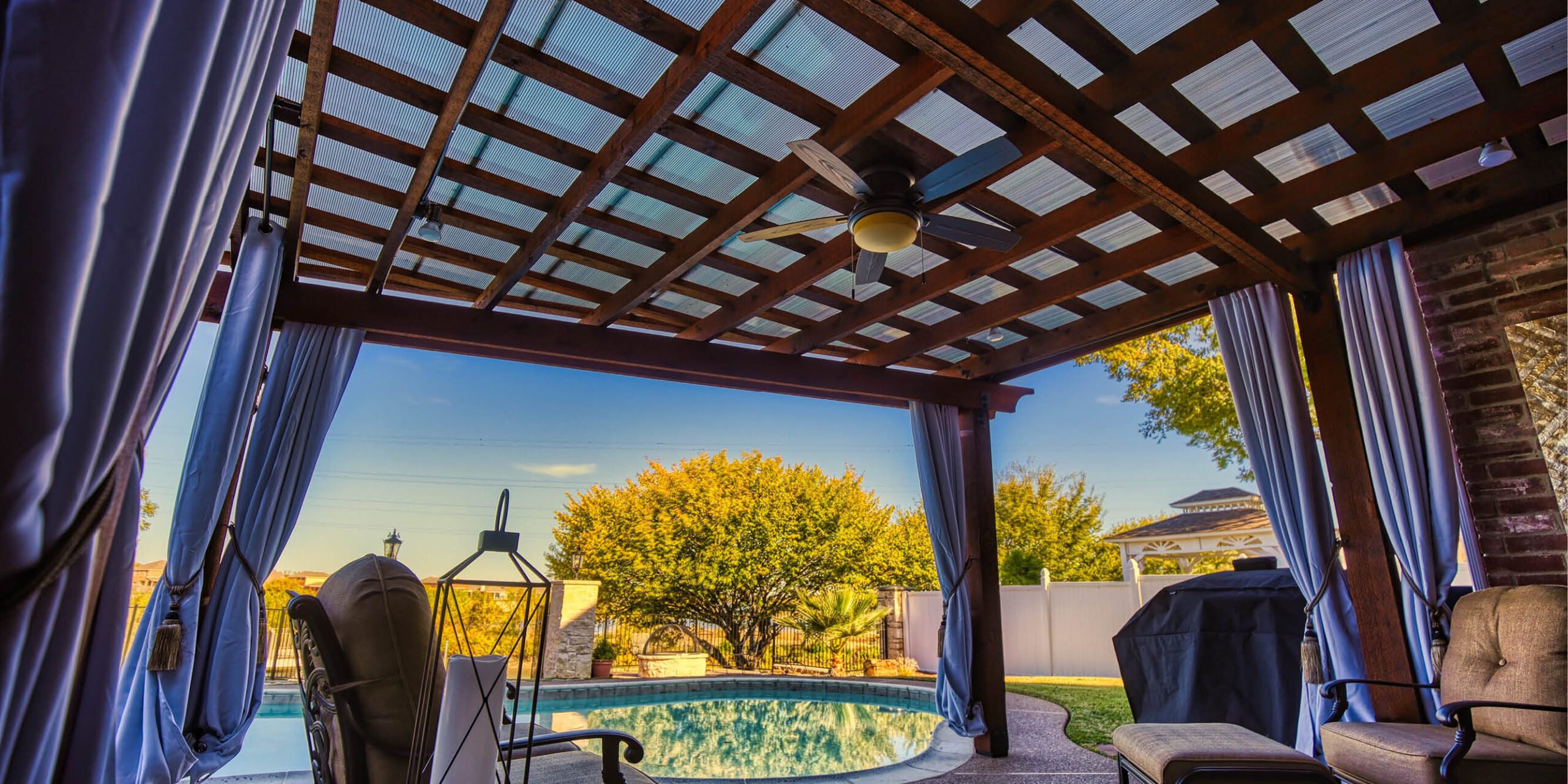 A covered patio area with drapery and a ceiling fan overlooking a swimming pool and landscaped garden at sunset