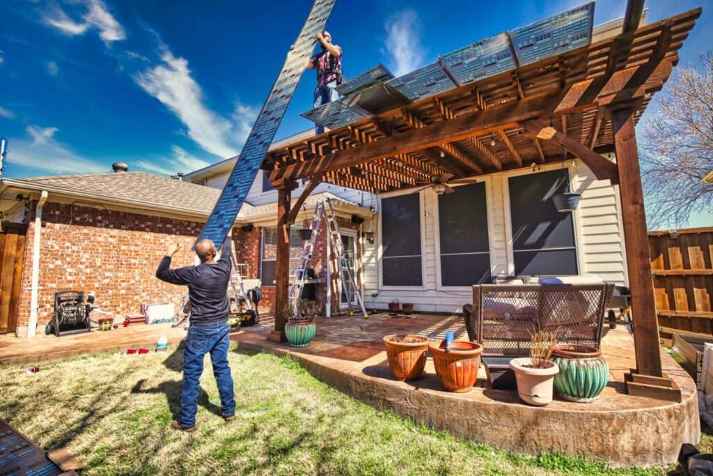 Two people installing solar panels on a wooden pergola in a sunny backyard with a brick house and potted plants.