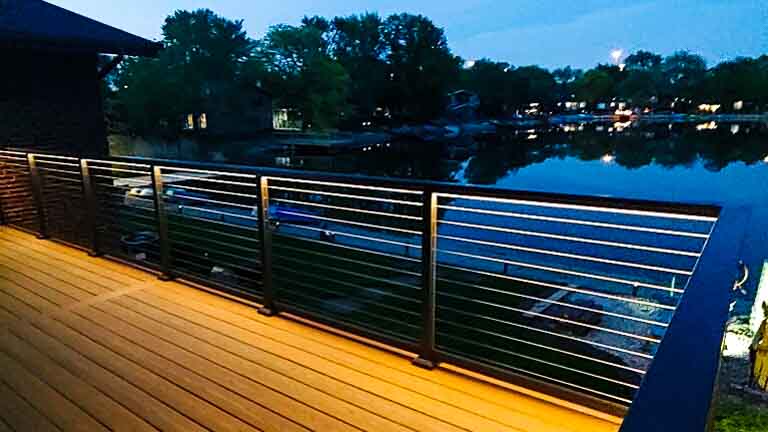 A waterfront deck with modern metal railings illuminated by soft deck lights, offering a view of a calm lake at dusk.