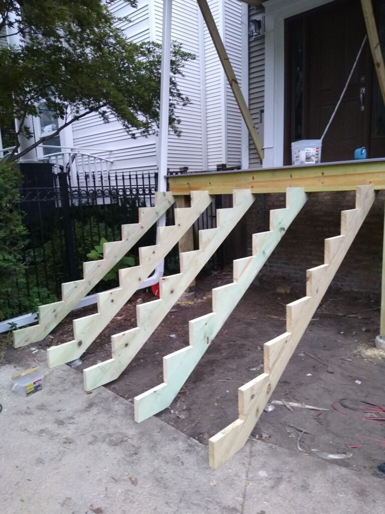 Unfinished wooden deck stairs in construction with tools and materials visible, adjacent to a house with white siding.