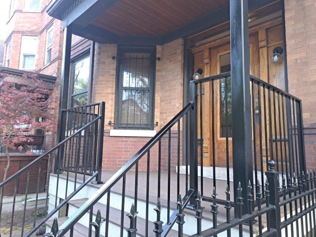Entrance of a brick building with wrought iron railings, wooden double doors, and a small porch with a protective overhang.