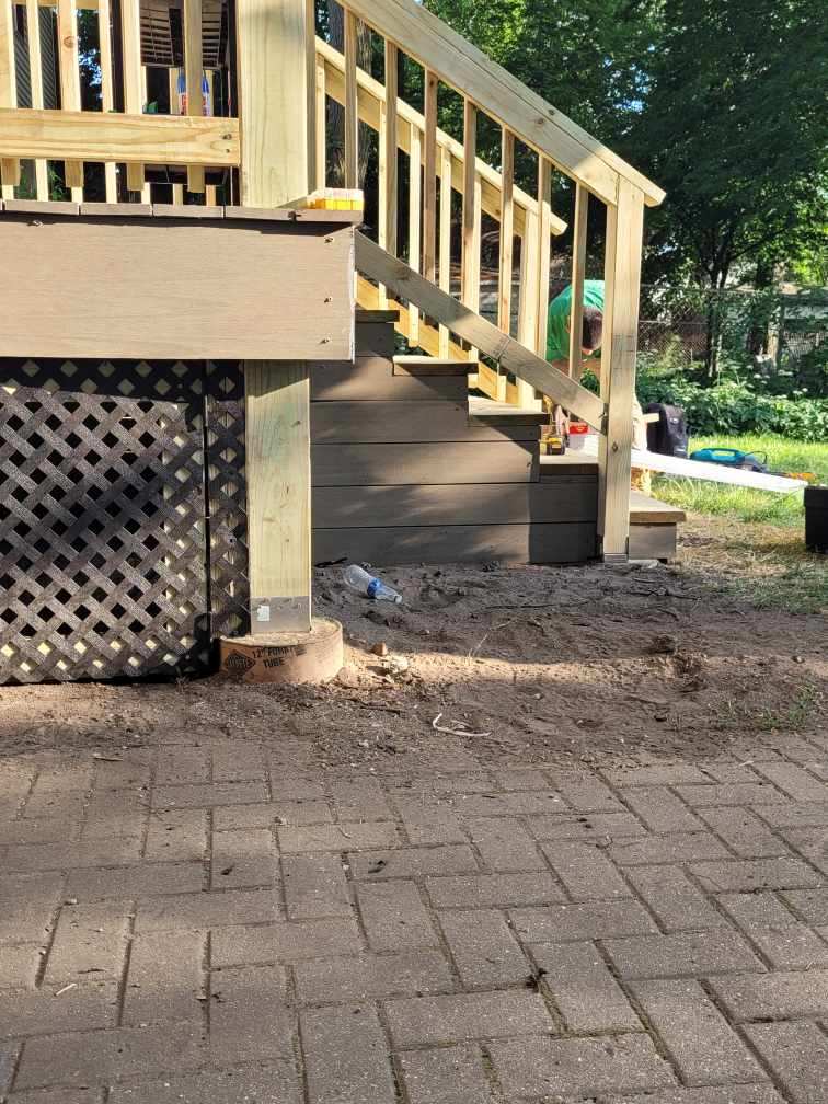 Partial view of a wooden deck staircase with lattice side panels overlooking a brick patio and a garden area in progress.-Porch Footing