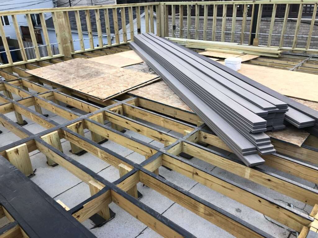 Construction materials including gray composite boards and plywood on a wooden deck structure in an outdoor setting.