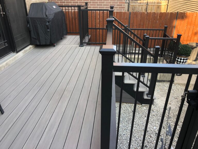 Composite decking with a black railing, outdoor furniture, and grill cover, adjoining a stone patio area.