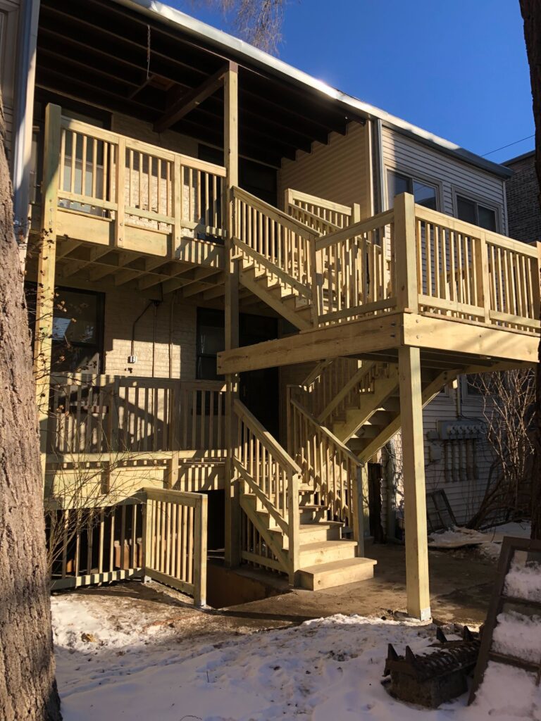 Sunlit wooden staircase and balcony attached to a house, with melting snow on the ground. (multi-level front porch)