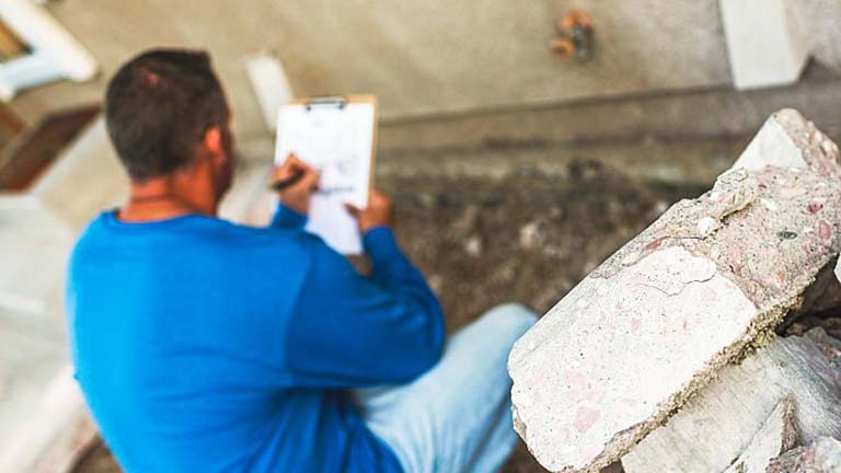 Inspector in blue shirt taking notes by a damaged concrete structure.