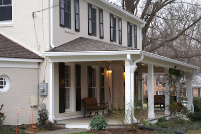 Charming front porch of a suburban home with cream siding, boasting white pillars and a solitary bench against the backdrop of mature trees