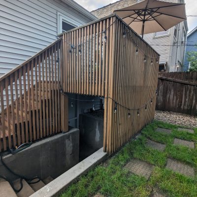 Wooden deck with vertical railing and an umbrella, overlooking a basement entrance with concrete steps.-Porch Without Roof