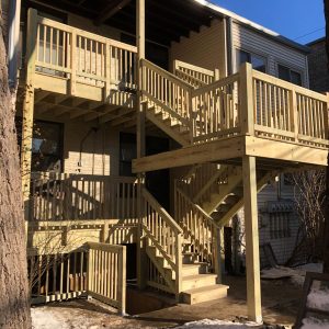 Sunlit wooden staircase and balcony attached to a house, with melting snow on the ground. (multi-level front porch)